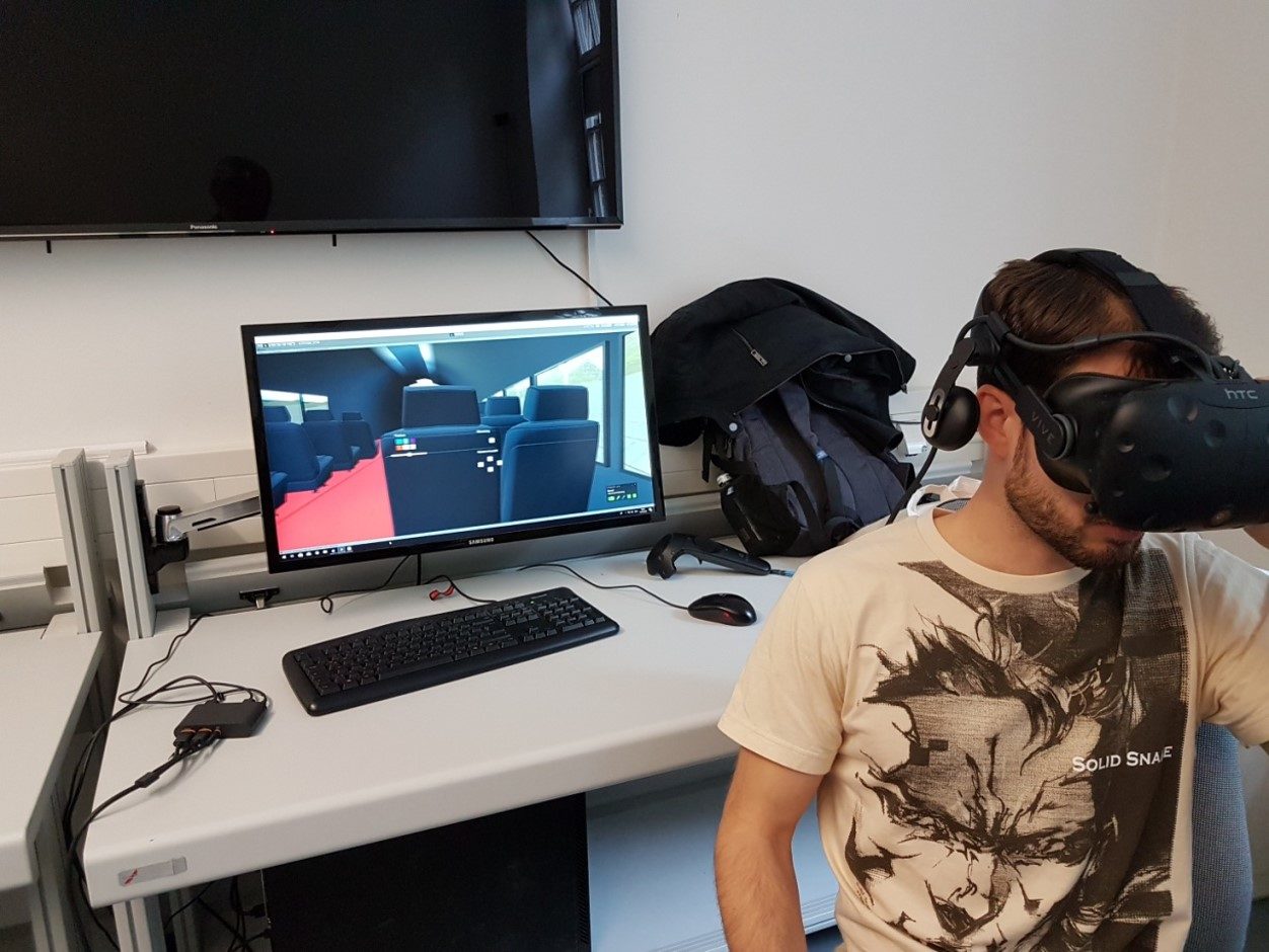 Foto von einem User, der eine VR-Brille trägt. Im Hintergrund sieht man auf einem PC-Bildschirm was dem User gerade angezeigt wird. Er befindet sich im Innenraum des Space-Liners und sitzt offenbar auf einem der Plätze, die an die Sitzreihen in einem Flugzeug erinnern.