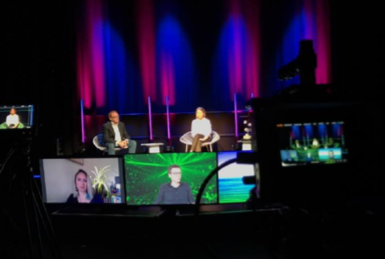 Foto einer Live-Veranstaltung, bei der zwei Menschen vor Ort auf einer Bühne sitzen. Um sie herum stehen große Bildschirme, auf denen vier andere Personen per Videochat zugeschaltet sind